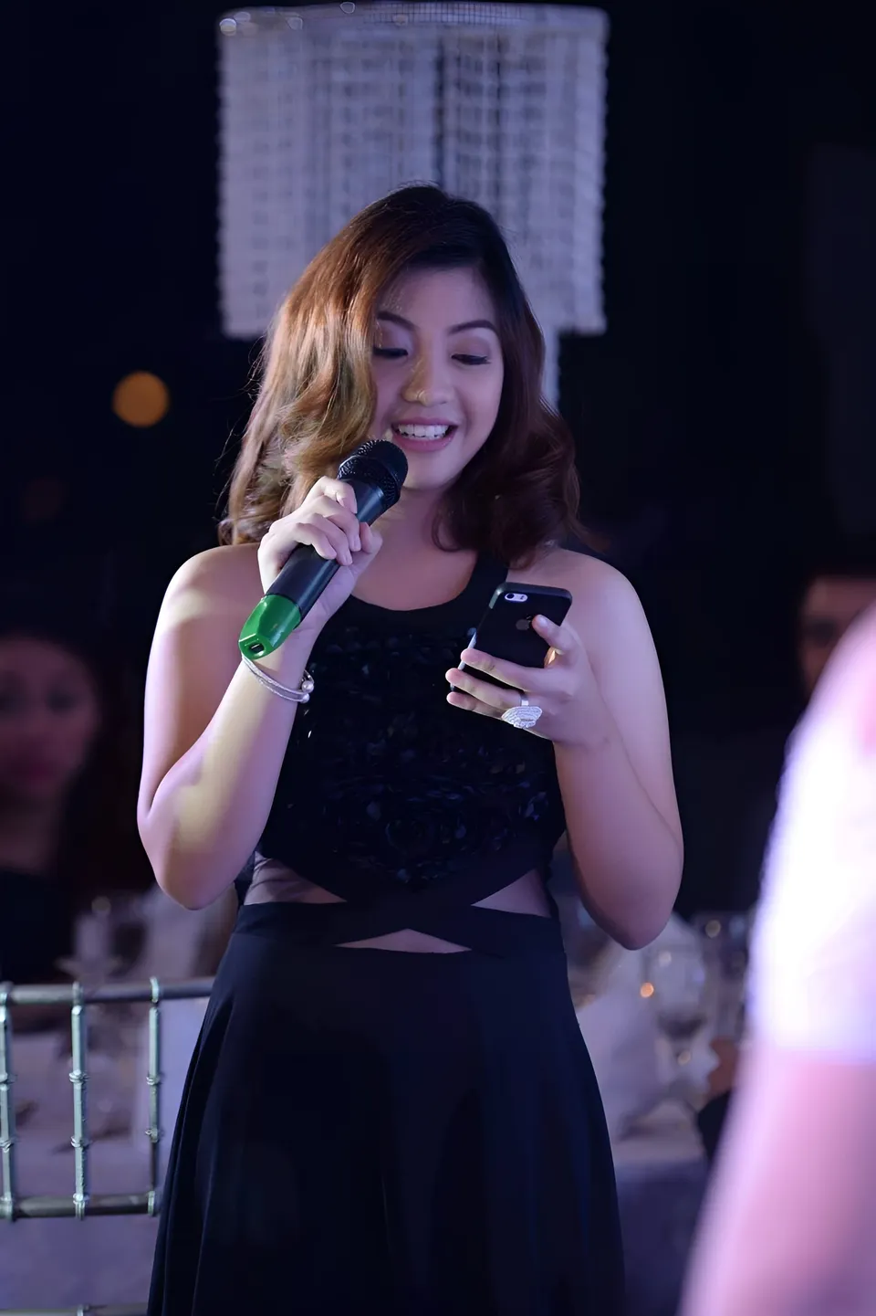Mariah Protacio in black dress holding microphone and phone while smiling