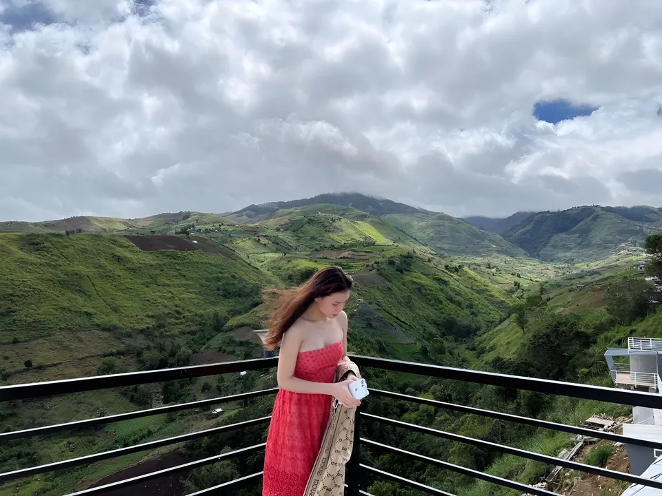 Marj Teves in red dress stands on balcony overlooking green hills