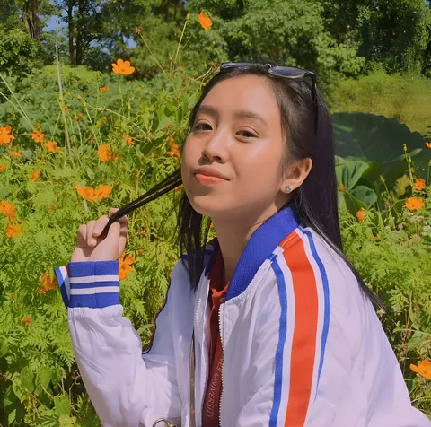 Marsell Cali wearing a striped jacket holds their hair outdoors among flowering plants