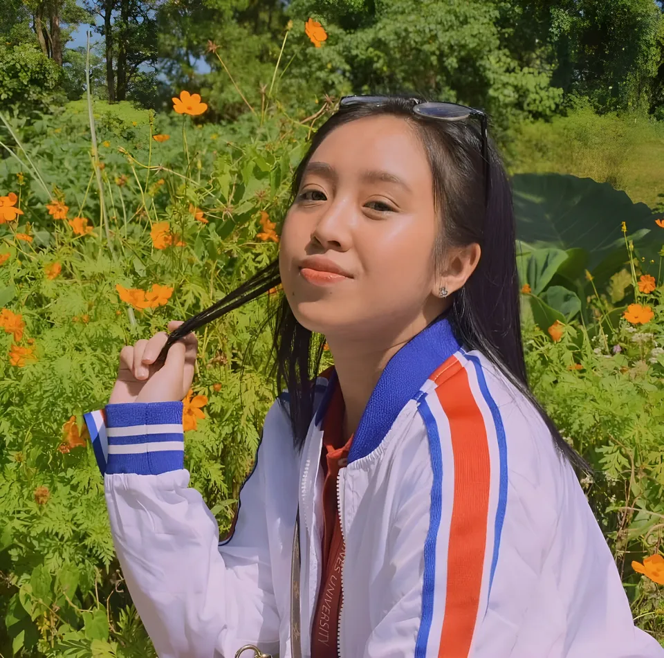 Marsell Cali wearing a striped jacket holds their hair outdoors among flowering plants