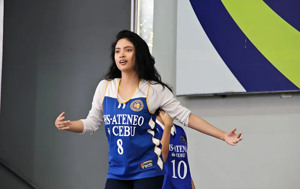 Mary Therese Rizon wearing a blue jersey over a gray top raises both arms indoors