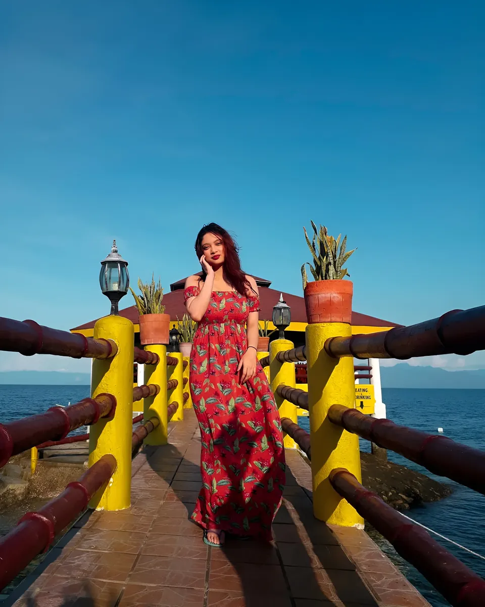 Mary Therese Rizon wearing a red floral dress poses on a wooden walkway beside the water