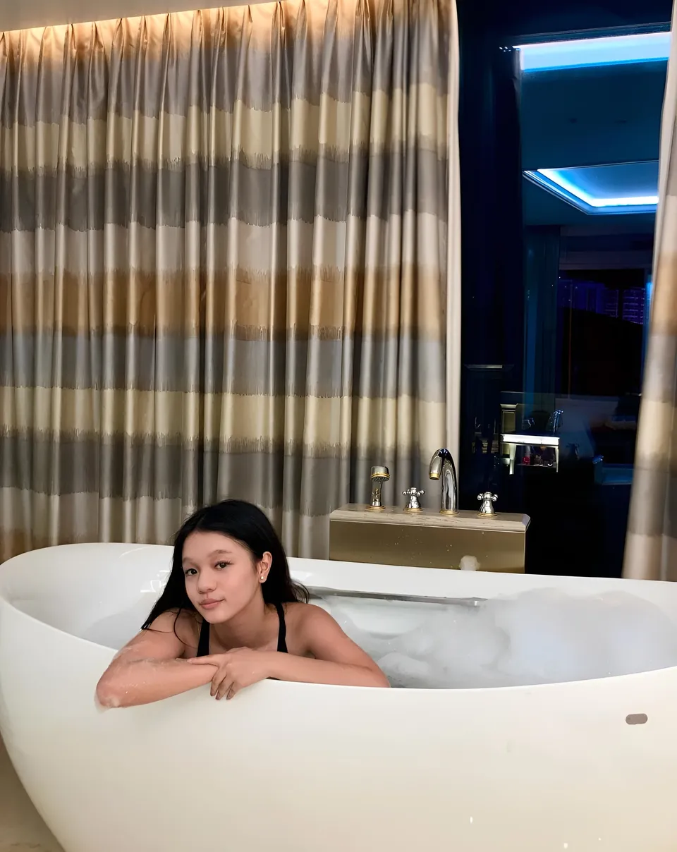 Maryshel Monroe in bathtub with foam, leaning on edge, behind patterned curtains