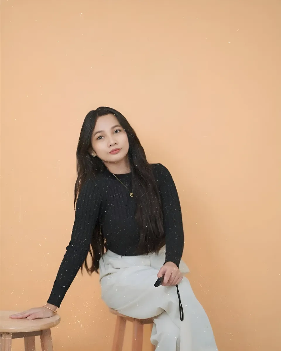 Mikay Alexis in black top and white pants seated on stool