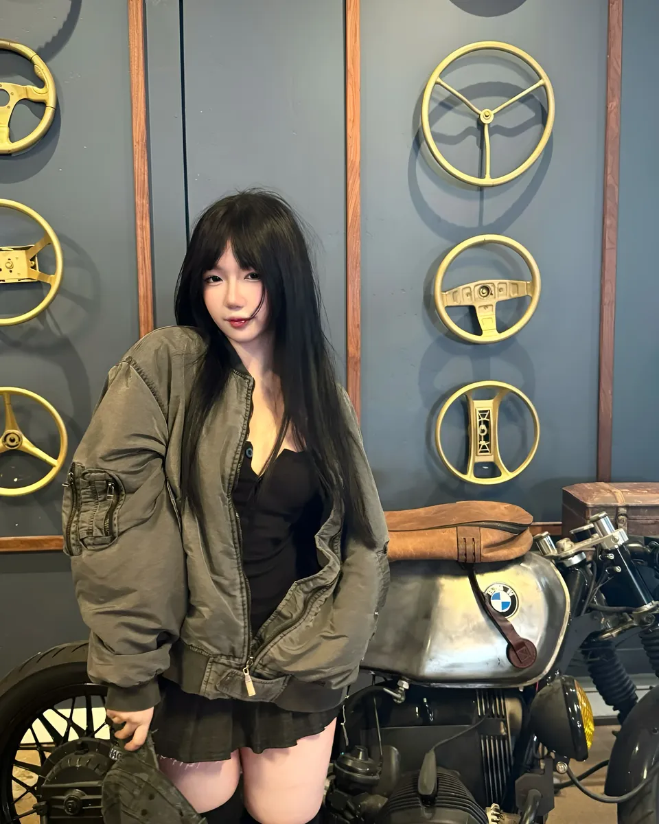 Mina Hartley in olive bomber jacket stands beside motorcycle with steering wheels on wall