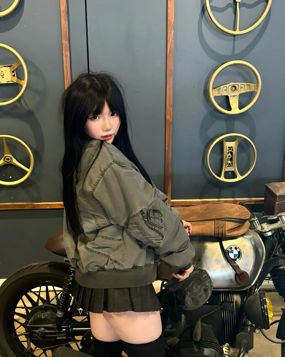 Mina Hartley in gray jacket seated on motorcycle beside wall with steering wheels