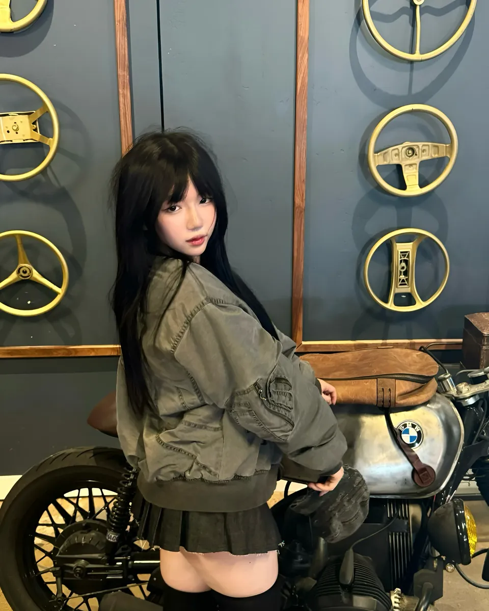 Mina Hartley in gray jacket seated on motorcycle with steering wheels on wall