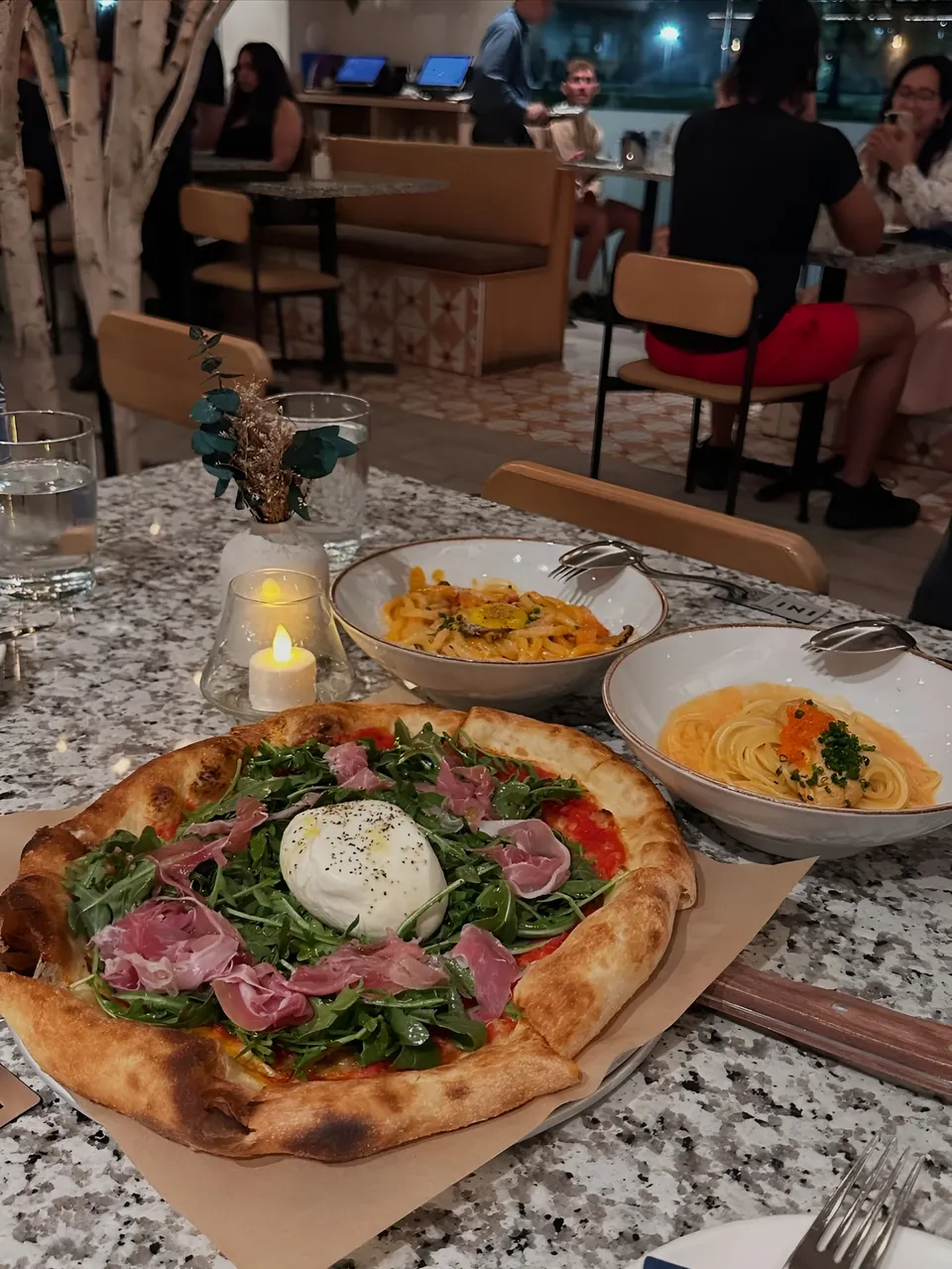 Mina Hartley — Table with pizza, pasta bowls, and candle in restaurant setting