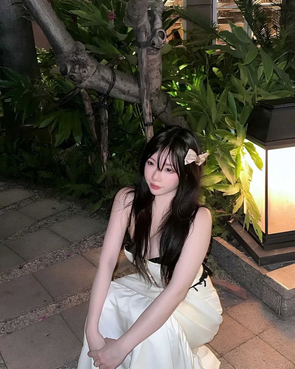 Mina Hartley in white dress seated outdoors near foliage and lantern