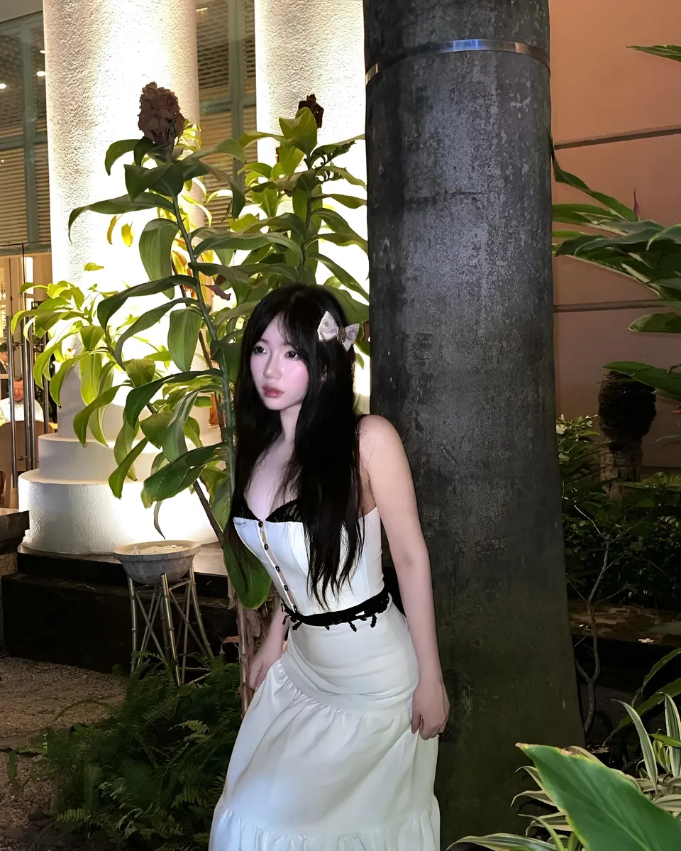 Mina Hartley in white dress standing beside pillar with plants