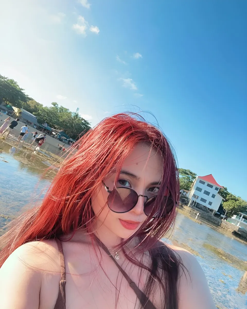 Mira Collins with red hair and sunglasses takes selfie at beach