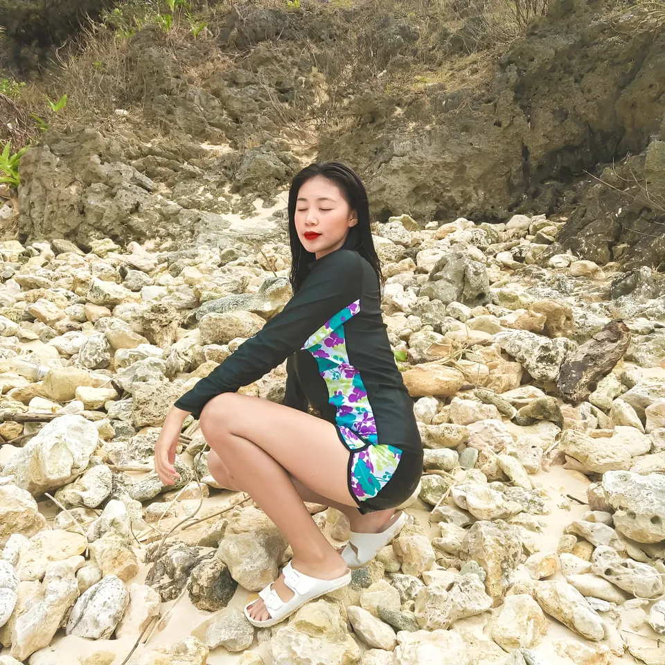 Moraya Ong in black and colorful swimsuit crouching on rocky beach