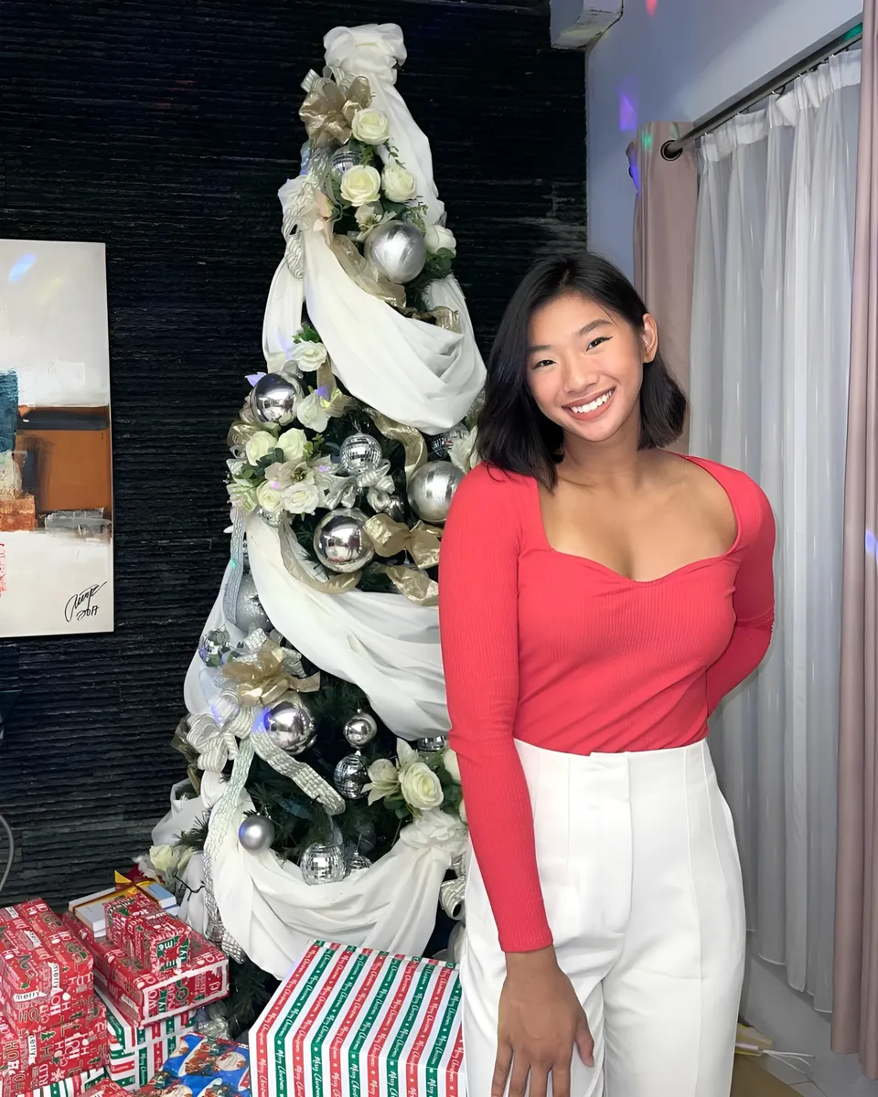 Pia Idelfonso wearing a red top and white pants poses beside a decorated tree