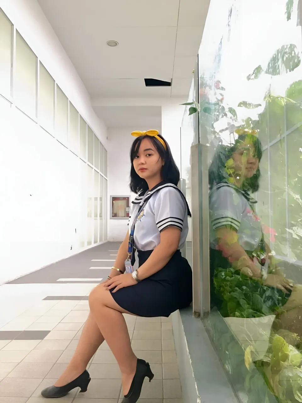 Sakura Keyema wearing a navy skirt and light top sits on a ledge in a windowed corridor