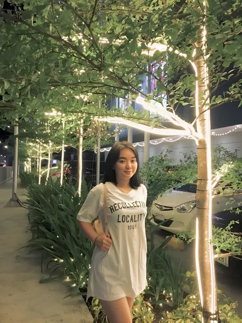 Sakura Keyema in white t-shirt dress standing under illuminated trees at night