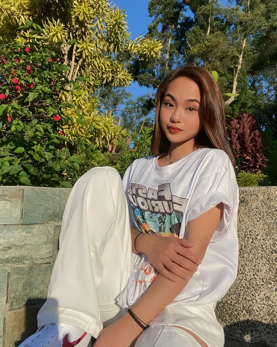 Shaira Adalyn in white top and pants sitting outdoors near plants