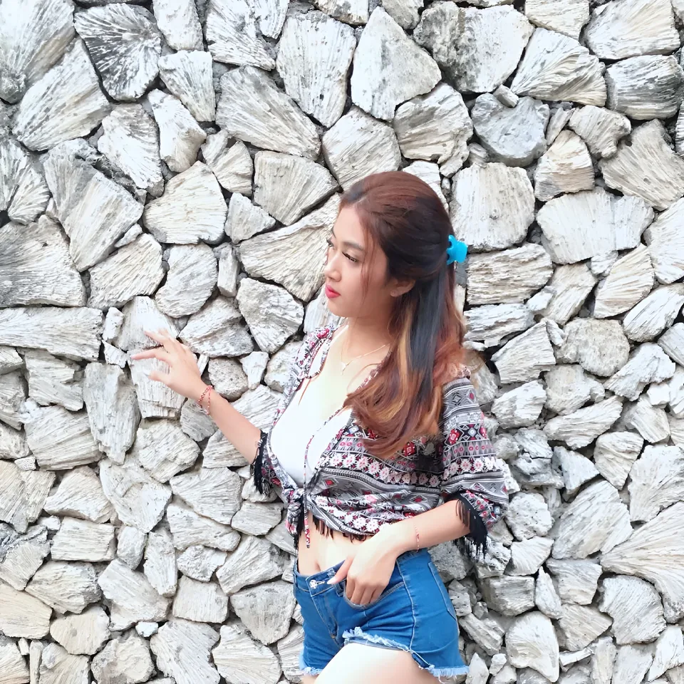 Sri Ayu Tarisa in patterned shirt and denim shorts stands against stone wall