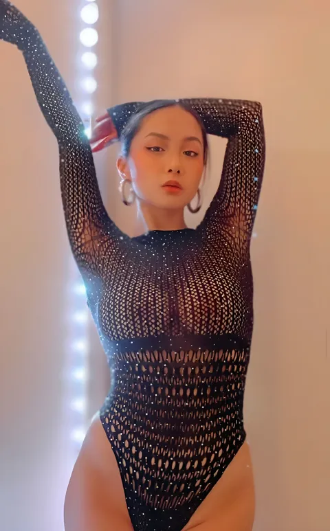 Stella Francesca wearing a black mesh bodysuit poses with both hands behind their head