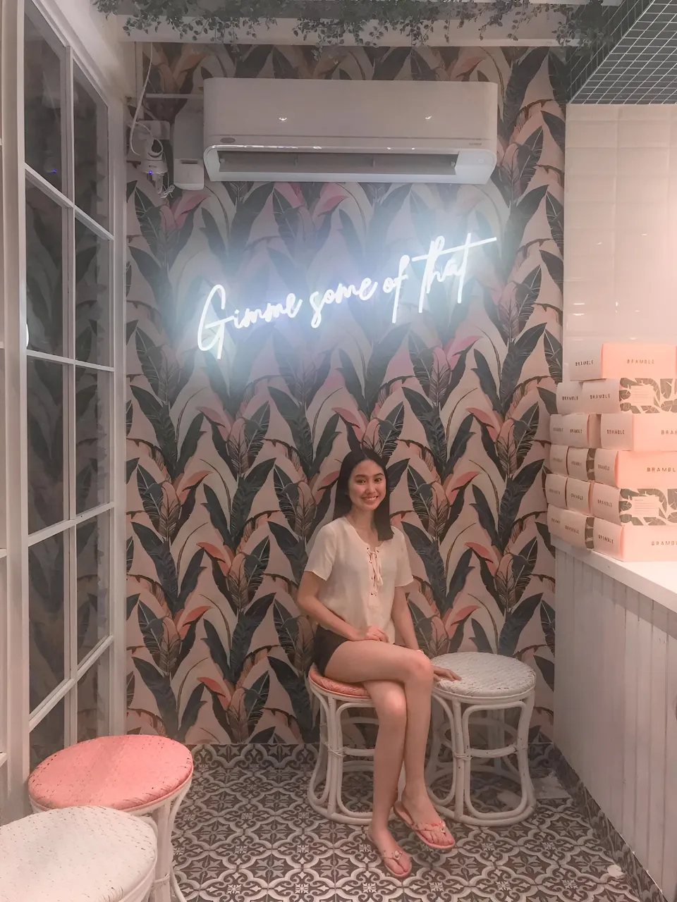 Stephanie Jean King sitting on stool in front of neon sign and tropical wallpaper