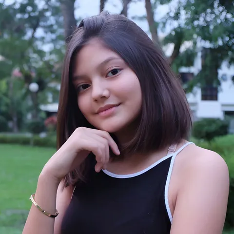 Via Gonzales wearing a black top with white trim rests one hand beneath their chin outdoors