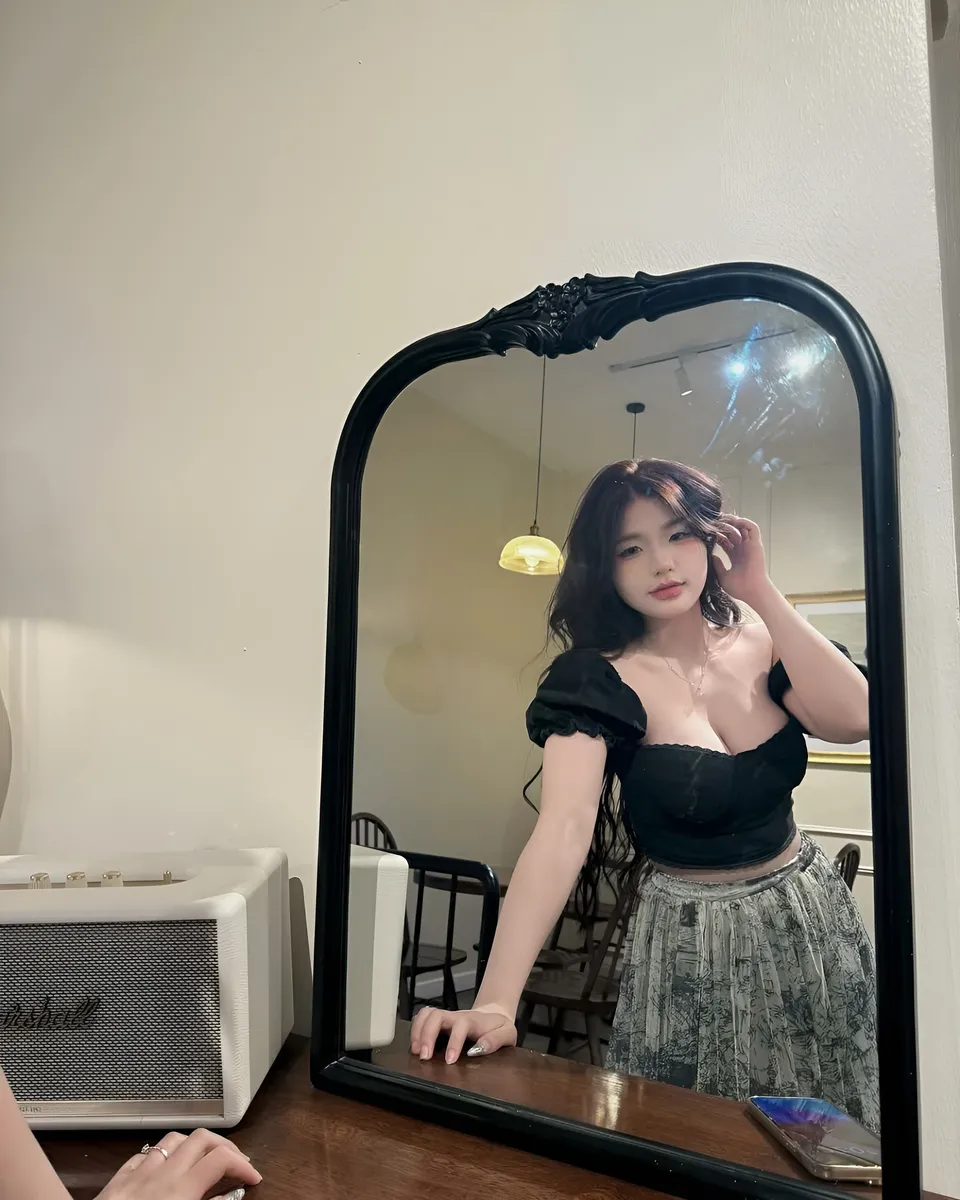 Yue Arden in black top and patterned skirt poses in front of mirror