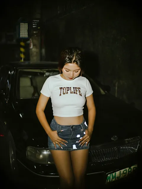 Zandra Hayes in white crop top and denim shorts stands beside black car