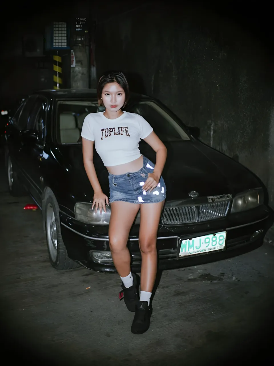 Zandra Hayes in white crop top and denim shorts stands beside black car