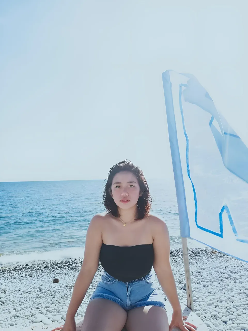 Zianne Tremedal in black top and denim shorts sits on pebble beach