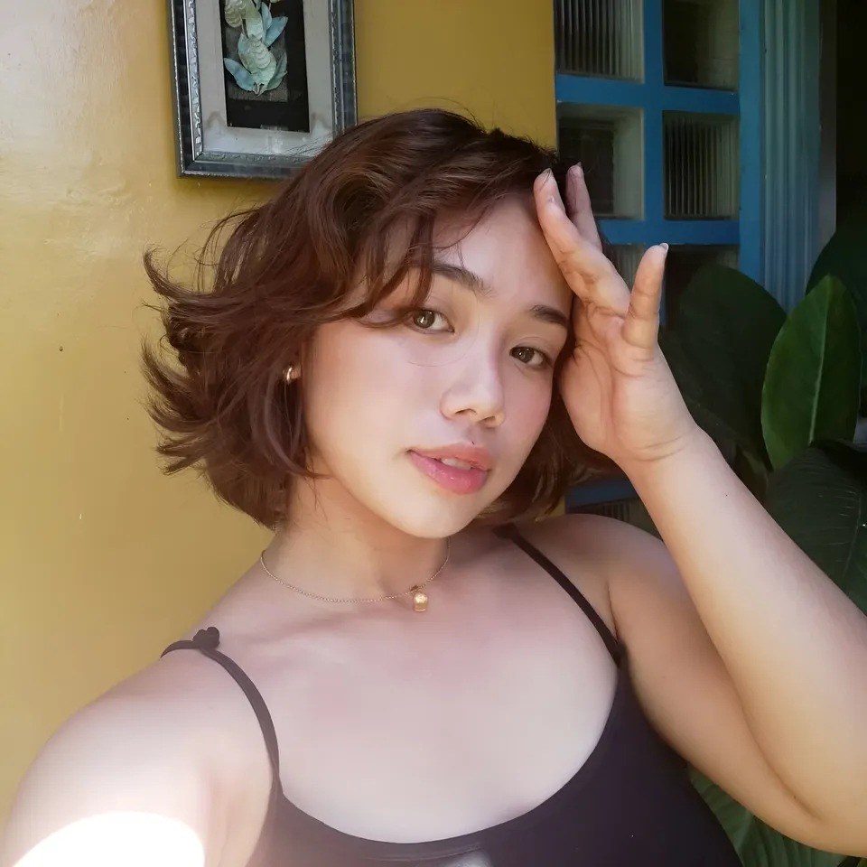 Zianne Tremedal in a brown top holds one hand against their forehead beside a yellow wall
